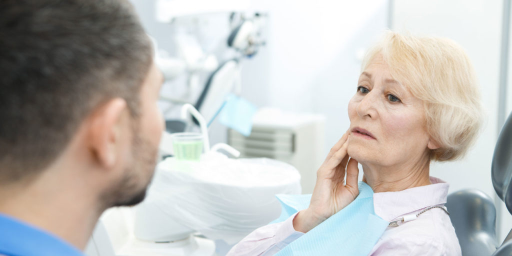 Elderly woman touching jaw in dental chair while discussing tooth pain with a dentist