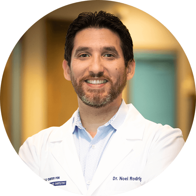Smiling portrait of Dr. Noel Rodriguez in a white coat, specialist in prosthodontics and dental implants